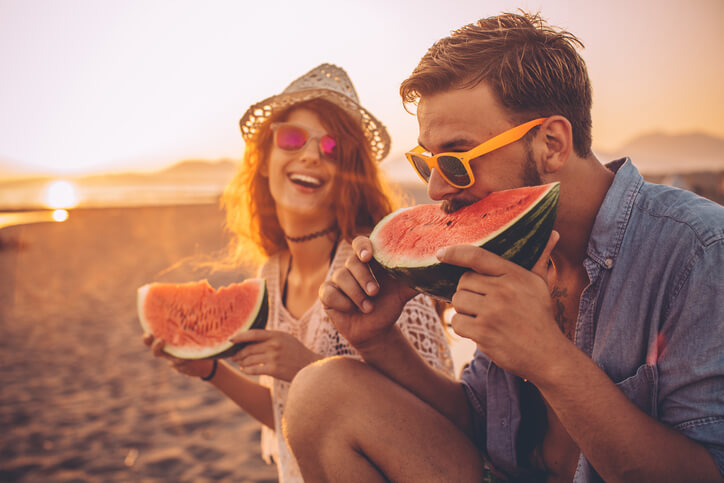 man and woman sitting on the beach eating watermelon